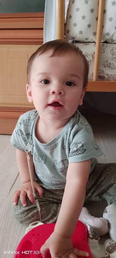 A young child, appearing to be around one to two years old, is sitting on a wooden floor. The child has fair skin, brown eyes, and short brown hair. They are wearing a light blue t-shirt with a pattern of construction vehicles and dark green pants with a leaf pattern. A clear plastic cord with a coiled spring is attached to the child's t-shirt, extending downwards towards an unseen object. The child is looking directly at the camera with an open mouth. In the background, a wooden crib with patterned bedding is visible, along with a section of a wooden cabinet. The lighting suggests an indoor setting, likely a home. The image is watermarked with "Infinix HOT 30i" in the lower left corner. The specific location of Kedrovy, Russia, cannot be determined from the visual information provided in the image.