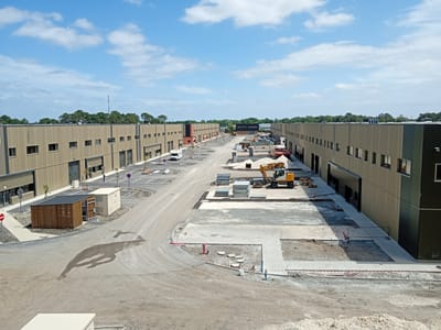 This elevated wide-angle image depicts a new commercial or industrial development under construction in Mérignac, France, under a clear blue sky with scattered cumulus clouds. The scene is brightly lit by direct sunlight.
The primary subjects are two long, rectilinear buildings with light beige/tan corrugated metal facades, each two stories tall, extending along the left and right sides of a central thoroughfare. These buildings feature multiple rectangular windows with dark frames on both levels and numerous dark grey roll-up garage doors or large entryways on the ground floor, indicating multiple units. The building on the left has a section towards its center with red-orange and dark grey paneling, providing a contrast to the uniform beige.
The central area consists predominantly of a wide, unpaved dirt road with visible tire tracks. Sections of this road and adjacent areas are undergoing paving or landscaping. Towards the left foreground, the dirt road curves around a newly paved section with white parking space markings, a wooden utility shed with a slatted facade and metal roof, and an adjacent cream-colored electrical box. A red circular stop sign and a circular no-entry sign on poles are visible near the paved area. A distinct, elongated, dark shadow of a person walking is cast on the dirt road, extending from the left side towards the center.
In the middle-ground and right-hand side, the central area remains largely unpaved and appears to be an active construction zone. A yellow wheeled excavator is parked near a large pile of sand and stacks of metal building panels. Various construction materials, including pipe sections, concrete covers, and barriers, are scattered across the ground. A red fire hydrant is visible on a small patch of poured concrete. Temporary black metal fencing marks certain boundaries.
The background beyond the buildings is characterized by a dense treeline of evergreen trees. In the far distance, above the treeline, a slender red and white communications tower is faintly visible. No individuals are explicitly visible within the scene. The overall environment indicates recent or ongoing commercial development.