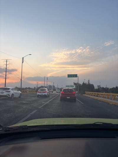 A highway scene during a sunset. Several cars are in traffic. A green highway sign is visible above the cars. The sky is colored with orange and yellow hues. The side of a vehicle is visible on the right side of the image, with a windshield wiper. Telephone poles are on the left.