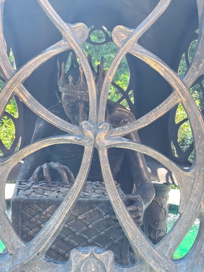 The image shows a bronze sculpture of a crowned figure hunched over a chest filled with coins. The sculpture is partially obscured by an ornate, circular metal grate, possibly part of a larger structure like a fountain or decorative fence. The figure wears a crown with intricate details and has a somber expression, with its head bowed and hands reaching into the pile of coins. The chest appears to be made of metal with a patterned surface. Behind the sculpture, lush green trees and foliage are visible, suggesting an outdoor setting. Sunlight is present, casting highlights on the metalwork and the sculpture. The water in the foreground, visible through the grate, is a bright turquoise color, further indicating a fountain or pool. The overall impression is one of a regal, perhaps greedy, figure guarding its treasure. No text is visible in the image. The location is identified as sovkhoza imeni Lenina, Russia.