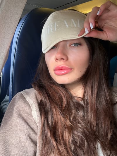 A young adult female is depicted in the cabin of an aircraft, likely within the vicinity of Lyon-Saint Exupéry Airport in Colombier-Saugnieu, France. She has long, wavy dark brown hair and fair skin. Her blue eyes are visible, and her lips are full with a glossy pink finish. She is wearing a light brown or beige ribbed sweater. Her right hand, featuring white manicured fingernails and a golden ring on the ring finger, is holding a light beige eye mask with the word "DREAM" printed on it, lifted to expose her right eye. Behind her, a dark blue aircraft seat is visible, confirming the interior setting of an airplane cabin. The left side of the frame shows a portion of the aircraft's interior wall or window structure. The subject is looking directly towards the camera.