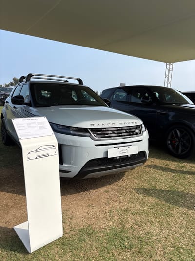 The image shows two Range Rover vehicles parked on a grassy area under a large canopy. The car in the foreground is a light-colored SUV, possibly white or silver, with a roof rack. In front of it, a white display stand with text and a sketch of an SUV is positioned. The Range Rover lettering is visible on the front grille. A dark-colored SUV, also a Range Rover, is parked to the right of the foreground vehicle. The environment appears to be outdoors, with dry grass and dirt on the ground. The sky is visible above the canopy. The scene suggests a car exhibition or dealership event. There are no people clearly visible in the frame. The location is described as the city of Lurín, Peru.