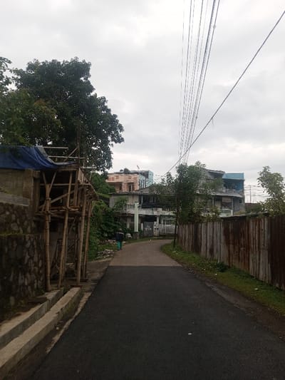 The image displays a residential street in Bhupalpally, India, under an overcast sky. A dark asphalt road curves gently to the left from the foreground. On the left side, a stone retaining wall with concrete steps borders a patch of dense vegetation and a structure under construction, featuring bamboo scaffolding and a blue tarp on its upper level. A single individual, wearing a dark top and light-colored trousers, walks away from the camera on the left edge of the road. To the right, a weathered, rusty corrugated metal fence runs parallel to the road, separated by a narrow strip of grass and debris. In the midground, visible beyond the road and vegetation, are multiple multi-story residential buildings. A prominent two-story building with a pale facade and balconies has "VOLOG" spray-painted on a ground-level door. Further back, another multi-story building with a light brown facade and rooftop scaffolding is discernible. Numerous overhead power lines traverse the upper right quadrant of the scene.