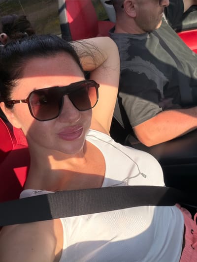 The image features two individuals inside a car, reportedly located in Matiushino, Russia. In the foreground, a woman with dark hair pulled back into a bun is capturing a selfie. She wears large, dark, aviator-style sunglasses with "MAYBACH" inscribed on the right lens. Her face shows freckles, and her lips have pink lipstick. She is dressed in a white ribbed tank top and a silver chain necklace with a small, stylized "B" pendant. A black seatbelt is diagonally across her chest. She is seated in a car with red leather upholstery visible on the seat and headrest behind her. Her arm is raised, resting on the seatback. In the background, slightly behind and to the right of the woman, a man is visible. He has short dark hair, a beard, and wears dark-tinted glasses. His dark-colored t-shirt has prominent dark, irregular wet patches on the front. He is seated in a dark car seat with red stitching, his head turned slightly away from the camera's direct line of sight. Both individuals are illuminated by strong, warm, directional sunlight, casting shadows and highlights, consistent with an outdoor setting, possibly in an open-top vehicle.