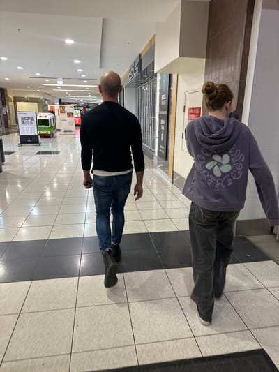 The image depicts the interior of a shopping center, likely located in Toormina, Australia, with a focus on the flooring and the backs of two individuals walking away from the camera. The floor is tiled with large square tiles, featuring a prominent alternating pattern of light beige and dark grey. The lighting appears to be artificial, typical of an indoor commercial space, casting reflections on the polished floor.
In the foreground, the lower legs and feet of two people are visible. The person on the left is wearing dark denim jeans and black boots, with the lower portion of their left leg prominently featured. The person on the right is wearing dark grey jeans and appears to be wearing sneakers.
The mid-ground reveals more of the individuals. The person on the left has a shaved head and is wearing a black long-sleeved shirt and blue jeans. The person on the right is wearing a purple hooded sweatshirt with a floral graphic design on the back, their hair is pulled up in a bun.
The background shows the architecture of the shopping center, including shopfronts with displays and signage. One sign reads "FIRE HOSE REEL." Further down the corridor, there are various retail displays and a small train-like ride. The environment suggests a public indoor space, possibly a mall or shopping precinct. The perspective is from behind the individuals, looking down the length of the corridor.