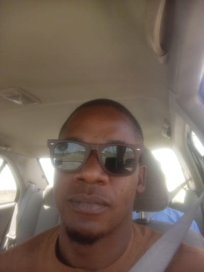A man is in the front seat of a car. He is wearing sunglasses and a seatbelt. The car's interior is beige. The reflection in his sunglasses shows a road. The location cannot be determined based solely on the image.