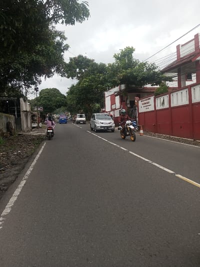 A street scene features a motorcycle approaching the camera, a silver car, and other vehicles further down the road. A woman on a scooter is on the left side of the road, heading in the opposite direction. Lush green trees line both sides of the street. A building with a red and white facade stands on the right, with a sign visible on its wall. The road is asphalt, with a yellow line on the right edge. The sky is overcast.