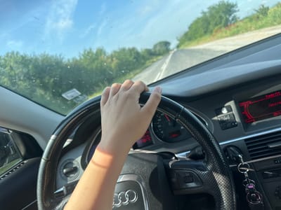 A first-person view from the driver's seat of an Audi vehicle shows a person's light-skinned hand gripping the top of a black, perforated leather steering wheel. The vehicle is in motion on an asphalt road in Ohoden, Bulgaria. The instrument cluster is visible behind the steering wheel, displaying a speed between 40-50 km/h, with "PRND S 6" and a "2650" reading, all illuminated with red backlighting. To the right, the car's infotainment screen shows "Radio FM" tuned to "108.0 MHz." A keychain with a purple lanyard hangs near the center console. The dirty windshield frames a clear blue sky with sparse white clouds above, and a two-lane road flanked by dense green trees and bushes stretching into the distance under bright sunlight.