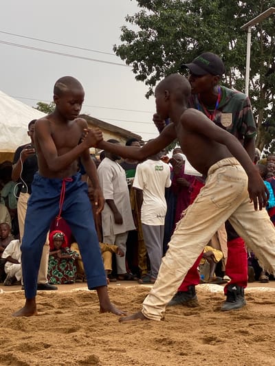 Two shirtless young men are wrestling in a sand pit in Kafanchan A, Nigeria. The wrestler closer to the viewer wears blue pants secured with a red drawstring and stands with his right foot firmly planted in the sand. His opponent is dressed in cream-colored pants and appears to be off-balance, poised on one foot. A referee wearing a camouflage shirt, black baseball cap, and lanyard stands above the wrestlers. Spectators surround the wrestling pit, including people dressed in traditional West African clothing. The event is held outdoors under an overcast sky. A few trees are visible.