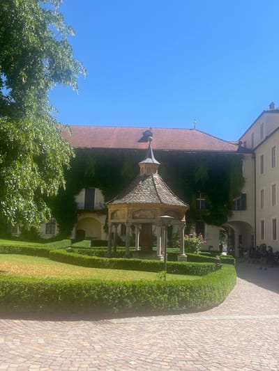 The image shows a courtyard with a stone pavement, manicured hedges, and a gazebo. The gazebo is octagonal with a tiled roof and decorative paintings on its sides. It is supported by columns. A lamppost stands to the right of the gazebo. The courtyard is surrounded by buildings with red tiled roofs. One building is covered in ivy, and another has a light-colored facade with multiple windows. In the background, several people are visible, some standing and some appearing to be near motorcycles. The sky is clear and blue. The scene appears to be a sunny day in a historical setting. The location is identified as Vahrn - Varna, Italy.