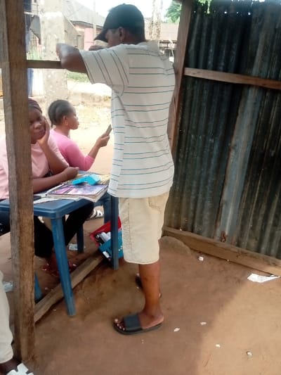 A man with a baseball cap, striped shirt, and khaki shorts leans against a wooden structure with corrugated metal. Two women sit at a table covered with magazines and a credit card reader. One woman rests her head on her hand, looking weary, while the other woman looks at her phone. A blue plastic crate lies on the ground beneath the table. The ground is dirt.