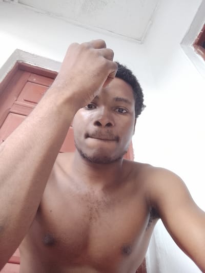 A shirtless young Black man is captured in a close-up, low-angle selfie taken outdoors. He has dark, curly hair and is looking directly at the camera with a neutral expression. His right arm is raised, with his hand resting on his forehead, obscuring part of his face. His chest and left arm are visible. The background features a white wall and a wooden door, suggesting an outdoor or semi-outdoor setting. The lighting appears bright, possibly indicating daytime. There is no visible text in the image. The overall context suggests a casual, personal photograph, possibly taken at home in Orodo, Nigeria.