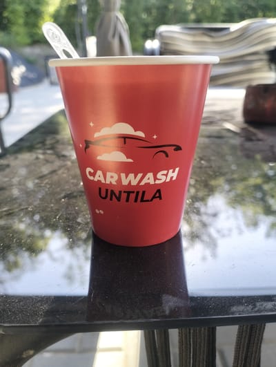 A close-up, eye-level shot shows a red paper cup with "CARWASH UNTILA" printed in white, along with a stylized image of a car and clouds. A clear plastic spoon protrudes from the cup. The cup rests on a reflective, dark surface, possibly a table, with visible scratches and dust. In the background, blurred greenery and outdoor furniture suggest an open-air setting. The image has a shallow depth of field, with the cup in sharp focus. The lighting is natural, indicating it was taken outdoors during the day. The scene is set in Șoldănești, Moldova.