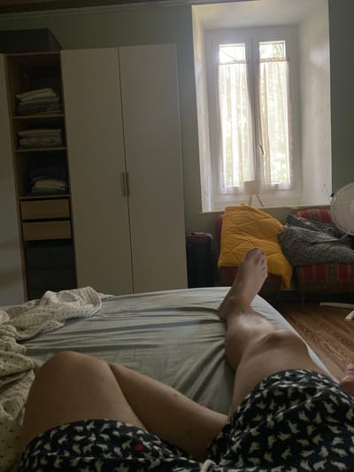 The image depicts a bedroom scene, likely within an apartment in Lonato del Garda, Italy. In the foreground, a person's legs are visible, reclining on a bed with blue and white striped sheets and a patterned duvet. The person is wearing dark blue shorts with a white elephant pattern. To the left, a tall white wardrobe with two doors stands against a wall. The wardrobe contains shelves with folded clothes and drawers. Beyond the bed, a window with sheer curtains allows natural light to enter the room, illuminating a small lamp on a wooden tripod and two chairs upholstered in patterned fabric. One chair is draped with a bright yellow quilted throw, and the other with a grey and floral patterned blanket. To the right of the chairs, a white oscillating fan is partially visible. The floor is made of wooden planks. The walls are painted a light green color. The overall impression is that of a domestic setting, possibly a bedroom in a residence or a hotel.