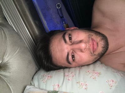 A light-skinned man with dark hair and a beard is lying on a floral pillow. His eyes are open, and he gazes at the camera. Part of his bare shoulder is visible. Behind him is a pleated curtain and a wall, illuminated with a blue light, and a hanging decorative object. A couch is to the left of the subject.