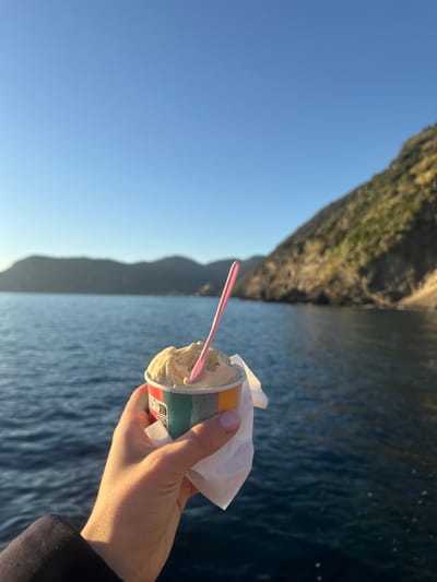 A first-person perspective photograph captured from Vernazza, Italy, depicts a human hand holding a cup of gelato against the backdrop of the Ligurian Sea and the rocky coastline. The hand, visible from the wrist with manicured fingernails painted light pink, holds a white paper cup containing light yellow gelato. A small, light pink plastic spoon is inserted into the gelato. The cup features vertical stripes of red, orange, yellow, green, and blue, partially obscured by a white paper napkin wrapped around it. The background consists of the deep blue, subtly rippled sea extending to distant, steep, vegetated cliffs under a clear, bright blue sky. Golden hour sunlight, likely from the late afternoon, illuminates the scene from the right, highlighting the gelato, the hand, and portions of the distant cliffs.