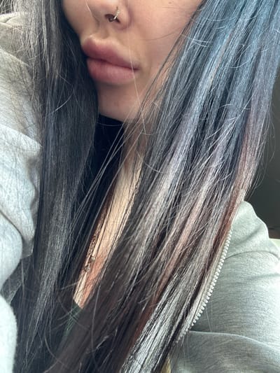 The image shows a close-up, head-and-shoulders shot of a person's face and upper body, with a focus on their lips, nose, and dark hair. The person is wearing a silver nose ring and a simple necklace. They are dressed in a grey hooded jacket. The background is blurred, but suggests an indoor setting. The image does not contain any information about the city of Monastyrshchina, Russia.