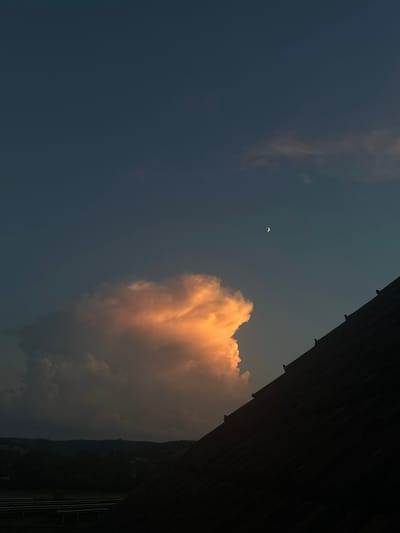 The image depicts a twilight sky over Heinebach, Germany. A large, illuminated cumulus cloud, lit orange by the setting sun, dominates the left side of the frame. A sliver of the crescent moon is visible in the upper right quadrant of the sky. In the foreground, the dark silhouette of a steeply pitched roof, possibly of a residential building, extends diagonally from the bottom right corner upwards. The roof appears to have several small, dark objects resembling birds perched on its surface. Below the cloud and to the left, a dark, silhouetted landscape of trees and rolling hills can be seen, with a hint of what might be solar panels or agricultural infrastructure in the far left foreground. The overall mood is serene and atmospheric, characteristic of the transition from day to night.