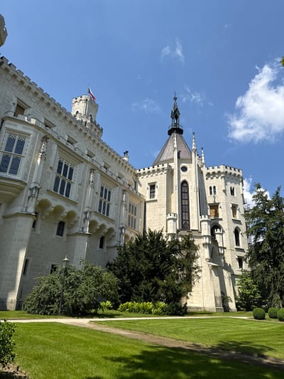 The image shows Hluboká Castle, a Neo-Gothic style castle located in Hluboká nad Vltavou, Czechia. The castle is made of white stone and features several towers, spires, and intricate architectural details. A Czech flag is visible on one of the towers. The castle is surrounded by a lush green lawn, manicured hedges, and mature trees. A lamppost stands on the lawn near the castle. The sky is blue with scattered white clouds. The image appears to be taken on a sunny day. There are no people visible in the image.