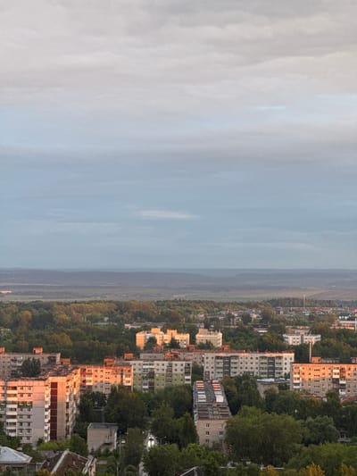 This image depicts an aerial or elevated view of a residential area in Kitsjanovo, Russia, under an overcast sky during a low-sun period, likely dawn or dusk. The upper two-thirds of the frame are dominated by a layered sky, transitioning from light grey at the zenith to a muted blue-grey near the horizon, indicating atmospheric haze and extensive cloud cover.

In the distant background, a faint line of dark, undulating hills or low mountains is visible, appearing dark blue-grey due to distance and atmospheric conditions. Below this, a broad band of dark green and muted brown landscape, comprising forests and possibly agricultural fields, stretches across the mid-ground.

The lower half of the image showcases an urban environment densely integrated with greenery. Multi-story residential buildings, primarily 5-9 story apartment blocks characteristic of Soviet-era architecture (e.g., Khrushchyovkas or Brezhnevkas), are the dominant structures. Several buildings on the left and center-right feature facades illuminated by a warm, golden-orange light, indicating a low sun angle from the left side of the frame. These buildings exhibit concrete panel construction, multiple windows, and balconies, some enclosed. Other buildings appear in shades of light grey, beige, or pale green. One lighter-colored building in the upper-right mid-ground features a more ornate architectural style with white facades and arched upper windows.

Dense clusters of deciduous trees, in various shades of green and some with yellowish autumnal hues, are interspersed throughout the residential area, partially obscuring buildings and roads. A winding asphalt road is visible in the lower center-right, with a vehicle featuring illuminated headlights, suggesting low light conditions. A tall, slender pole, possibly a communication mast or utility pole, stands near the center-right. No individuals are visibly present or engaged in specific actions within the frame.