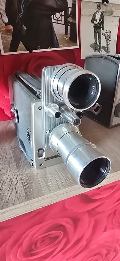 The image shows a vintage film camera on a wooden surface. The camera is silver and has a large lens. Behind the camera, there are two framed photographs. The photograph on the left shows a person in dark clothing. The photograph on the right shows a man and a child. The background is a red fabric with a rose pattern. This scene is located in the city of Gostagayevskaya, Russia.