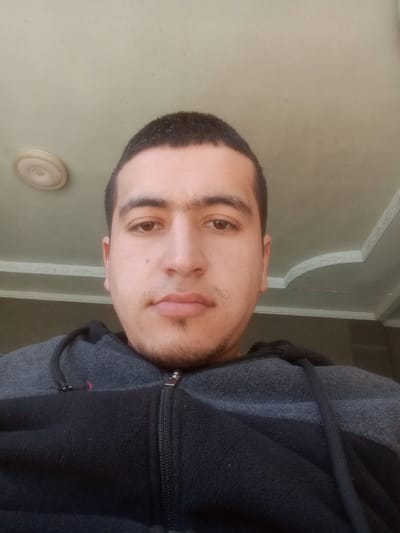 A close-up, head-and-shoulders portrait of a single male subject, facing forward and looking directly at the viewer, taken indoors in Kasan, Uzbekistan. The subject possesses short, dark hair, dark eyebrows, brown eyes, and light facial stubble on the chin and upper lip. He is attired in a two-tone fleece jacket or hoodie featuring a gray upper section and a black lower section, with a central zipper and dark drawstrings visible at the neckline. The background consists of a light-colored ceiling. A round, recessed light fixture with a decorative rim is positioned in the upper-left quadrant of the frame, and white decorative ceiling molding is visible along the upper-right edge.