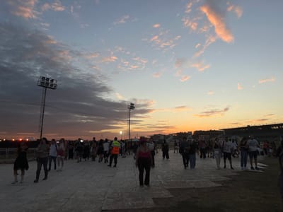 The image displays an outdoor event venue in Martínez, Argentina, at dusk. The sky presents a gradient of color, transitioning from warm orange and pink hues near the horizon to light blue and grey overhead, with scattered, textured clouds. The ground in the foreground is covered with temporary, light-colored modular panels, extending into the midground, with areas of darker ground (likely grass or dirt) visible on the right.
Numerous individuals, predominantly adults, are present throughout the midground, generally walking from left to right or standing. Their attire is casual and varied. A male individual in an orange high-visibility vest is visible in the left-center midground, facing right. Two prominent, tall metal floodlight towers with multiple lamp fixtures are visible; one on the far left and another in the middle-right distance.
In the far background on the right, a multi-tiered structure, consistent with grandstand seating, is silhouetted against the horizon, indicative of a large outdoor stadium or racetrack. Distant artificial lights are visible on the left horizon. The overall lighting is dimming natural light from the setting sun, supplemented by nascent artificial illumination.