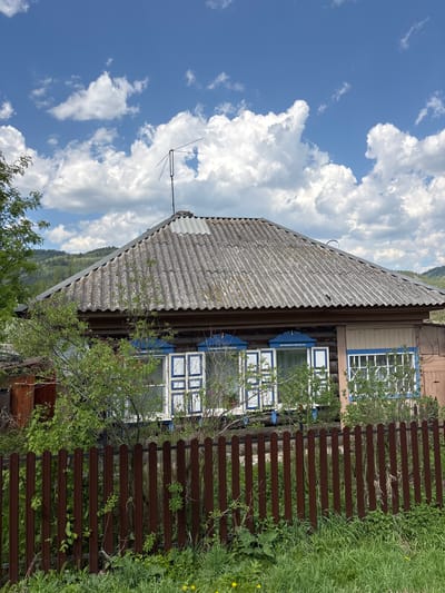 The image depicts a single-story wooden house with a gray asbestos roof and blue-trimmed windows. An antenna extends from the roof toward the sky, filled with fluffy white clouds. A brown wooden fence stands along the side of the house, partly obscured by vegetation and tall grass. The house is located in Овсянка, Russia.