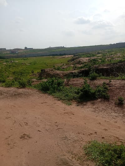 This is an outdoor, eye-level, medium shot photograph of a landscape with construction in the distance. In the foreground, a dirt path is visible with various footprints in the sand. Lush green grass grows along the edges of the path and in other areas of the foreground. The middle ground features uneven terrain with some exposed earth and more green vegetation. In the background, a long concrete wall stretches horizontally across the frame, marking the boundary of a construction site. Beyond the wall, there are buildings under construction, including what appear to be shipping containers repurposed as temporary structures. The sky is mostly overcast with scattered clouds. The location is Karsana 2, Nigeria, indicated by the presence of the construction site and associated infrastructure. There are no people or animals visible in the image.