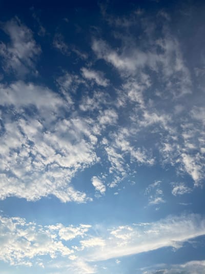 The image captures a broad view of a bright blue sky filled with scattered white clouds. The clouds vary in density and form, with some appearing wispy and thin, while others are more puffy and voluminous. Sunlight filters through the clouds, creating variations in brightness across the sky. The composition suggests an outdoor setting, likely during daytime, given the clear blue sky and bright illumination. There are no people, objects, or text visible in the image. The scene evokes a sense of openness and natural beauty, typical of a clear day. The orientation of the image is such that the sky fills the entire frame, with no ground or horizon visible. The clouds are spread across the frame, with a notable concentration of brighter, more illuminated clouds towards the right side, suggesting the direction of sunlight. The overall impression is one of a calm and pleasant atmosphere.