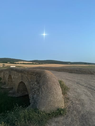 The image depicts an ancient stone aqueduct archway, likely in Filipeni, Moldova. The aqueduct is constructed from rough-hewn stone blocks, with visible signs of age and erosion. Lush green grass and small wildflowers grow around and within the structure, suggesting a natural setting and possibly an extended period of disuse. The scene is bathed in the bright light of a clear, blue sky, with a distinct starburst lens flare from the sun indicating it is likely midday. Beyond the aqueduct, the landscape unfolds into rolling hills covered with fields of dry, golden grass and darker green, forested areas, creating a sense of rural expanse. The perspective is angled, looking across the top of the archway and into the distant landscape. There are no people or specific activities evident in the image, focusing the viewer's attention on the enduring structure and the natural environment. The overall impression is one of timelessness and natural beauty, with the ancient aqueduct serving as a silent monument within the tranquil countryside.