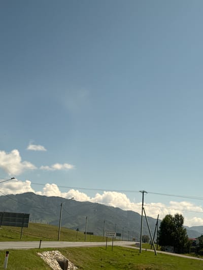 A wide shot captures a bright, sunny day in Tuekta, Russia. The sky is a clear, pale blue with scattered white clouds, stretching across the upper half of the frame. In the distance, rolling green hills rise up, their slopes covered in lush grass and dotted with trees. A paved road runs through the middle of the scene, curving slightly to the right. On the left side of the road, a large, grey sign with a grid pattern stands tall. Several streetlights are visible along the roadside, some illuminated, others not. A cluster of trees stands to the right of the road, with a few buildings partially visible behind them. A sign with Cyrillic lettering reads "КАРАКОЛ KARAKOL." The foreground is dominated by a grassy embankment with a culvert and scattered stones. No people are present in the image.