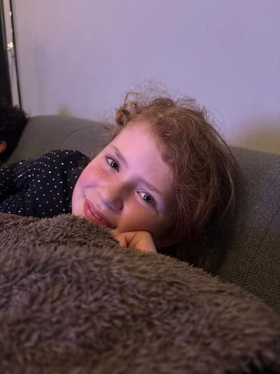 A young girl with red hair and freckles gazes upward with a slight smile. She rests her head on her hand, partially obscured by a fuzzy brown blanket. She wears a black top adorned with small silver dots. She seems to be lying down, with the head of another person visible in the bottom left corner. A white wall fills the upper left of the frame, contrasting with the darker hues surrounding the girl.
