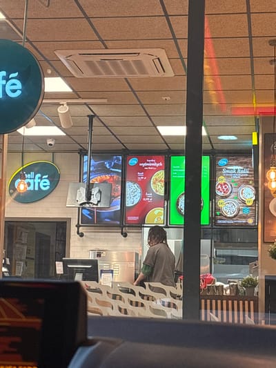 The image shows a reflection in a window, likely of a service station convenience store. The scene is slightly distorted due to the reflection.

Visible inside the store are several digital menu boards displaying food items. One board advertises "Smak Italii" (Taste of Italy) with images of pizzas. Another board shows "wyśmienitych zup Shell Cal" (delicious Shell Cal soups), with prices for "Rosół" and "Żurek". The "Shell Café" logo is also prominently displayed on a sign.

A person with dreadlocks, wearing a grey t-shirt, is partially visible inside, standing behind a counter. They appear to be interacting with something out of frame. There are also shelves with products and what looks like a small refrigerator or display unit visible in the background.

Outside, a modern ventilation unit is mounted on a tiled wall. The lighting suggests it could be daytime, but the reflection makes it difficult to determine the exact time or weather conditions. The overall impression is of a typical roadside service station offering food and drinks. The visible text, primarily in Polish, indicates promotional offerings for pizzas and soups at a Shell Café.
