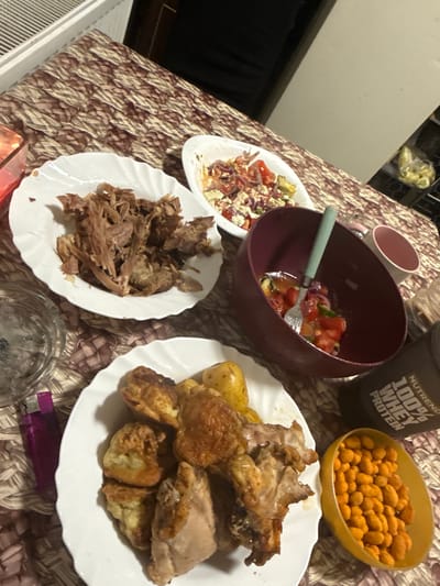 A low-angle, close-up shot captures a table laden with food. In the foreground, a white plate holds a portion of roasted chicken and what appear to be Yorkshire puddings, accompanied by small, roasted potatoes. To the right, a yellow bowl is filled with orange-coated peanuts. Behind these, a dark maroon bowl contains a chopped salad of tomatoes, red onion, and what looks like feta cheese, with a fork resting inside. A white bowl in the background also contains a similar salad. To the left, a pink lighter lies next to an ashtray. The table is covered with a patterned tablecloth. A black container with "NUTREND 100% WHEY PROTEIN" written on it is visible. The lighting is dim, suggesting an indoor setting, possibly a home dining area. There are no people visible in the image. The provided location of Jablonec nad Nisou, Czechia, is an external detail not discernible from the visual content.