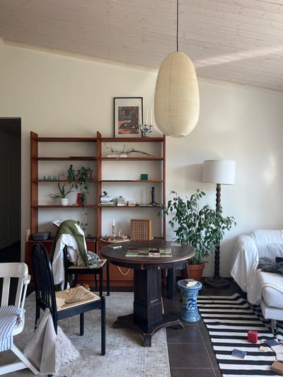 This is an indoor scene, likely a living room or dining area, featuring a collection of furniture and decorative items. The room has a sloped, wood-paneled ceiling.

In the center of the image, a dark wooden round table stands on a pedestal base. On the table are several tiles or coasters arranged in a pattern, and a few books. To the left of the table is a black wooden chair with a woven seat, draped with a green sweater and a white garment. To the left of this chair is a white chair with blue and white striped cushions.

Behind the table and chairs is a tall, wooden bookshelf filled with various items, including books, plants in pots, decorative objects, and a framed picture hanging on the wall above it. A large, cream-colored, lantern-style pendant light hangs from the ceiling above the table.

To the right of the table, a large green plant in a terracotta pot is placed next to a dark wooden floor lamp with a white shade. A portion of a white sofa is visible to the right of the lamp, covered with a textured throw. A blue and white ceramic garden stool stands near the table.

The floor is a combination of dark tiles and a patterned rug with black and white stripes. Scattered on the rug are several small objects, including a red gift box.

There are no people visible in the image. The scene appears to be a still life arrangement within a domestic setting. The location is identified as Hämeenlinna, Finland.