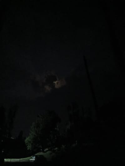 A nighttime outdoor shot captured a dimly lit scene dominated by a dark, cloudy sky. A faint illumination from the moon is visible through a break in the clouds, casting a diffused glow. Silhouetted trees and vegetation are present in the foreground and midground, creating a shadowy landscape. A dark, indistinct object, possibly a vehicle, is visible in the lower left portion of the frame. No people are present. The location is identified as Auk, Kyrgyzstan.