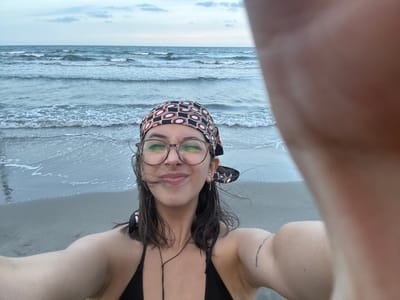 The image shows a person on a beach, likely in Mata de Uva, Mexico, taking a selfie. The person is wearing a black bathing suit and a patterned headscarf. They also have glasses and appear to have a tattoo on their arm. The background includes the ocean and the sky. The person is facing downward and their arm is partially covering the lens.