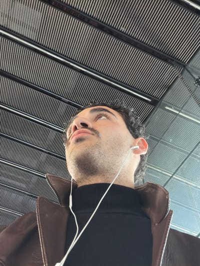A low-angle, close-up photograph depicts an adult male in an indoor public setting within Mérignac, France, consistent with Bordeaux–Mérignac Airport. The subject, visible from the upper chest, has dark, curly hair, a trimmed mustache, and stubble, and is wearing a dark turtleneck beneath a brown leather jacket. White wired earphones are inserted into both ears, with the cables extending downwards. The male's gaze is directed upwards and slightly to his left. The overhead environment features a structured ceiling composed of multiple parallel dark beams interspersed with lighter, finely-ribbed panels, extending diagonally across the upper frame.