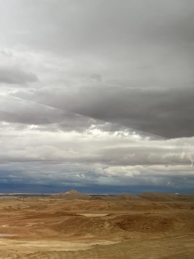 The image depicts a vast, arid desert landscape under a heavily clouded sky within the Arava Tichona Regional Council, Israel. The upper two-thirds of the frame are filled with a complex cloud formation, featuring layers of white, light grey, and dark grey stratus and cumulus clouds, with darker, thicker storm clouds dominating the upper section and creating an overcast condition. Faint diagonal light reflections are visible across the sky portion, suggesting the photograph was taken through glass. The lower third of the image shows an undulating terrain composed of reddish-brown and tan soil, indicative of a barren desert environment. Rolling hills and depressions characterize the foreground and mid-ground. In the mid-ground, several thin, dark vertical structures, resembling utility poles or communication masts, are faintly visible on the left. A small, light-colored patch of ground, possibly a dry or damp depression, is present slightly left of center in the mid-ground. The distant horizon reveals darker, muted hills or mountains beneath the cloudy sky. No human figures, animals, or significant man-made structures are present within the observable scene.