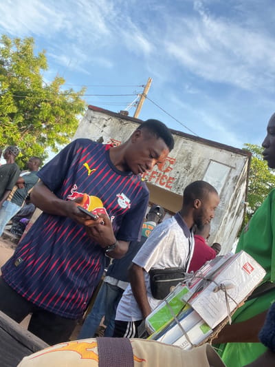 This image captures an outdoor street scene in Gbaza, Nigeria, featuring several individuals and architectural elements under a clear blue sky with scattered white clouds.

In the left foreground, a young Black male with short dark hair and a facial marking on his right cheek is depicted. He wears a navy blue Red Bull Leipzig soccer jersey with red vertical stripes and black shorts. Both hands hold a smartphone, which he is actively viewing. On his left wrist, a dark watch is visible.

To his right, a second young Black male with a short beard and dark hair wears a light grey polo shirt and a black fanny pack strapped across his chest. He is looking towards the right side of the frame.

On the far right, partially visible, a third Black male in a green shirt is present. He carries a stack of cardboard boxes, appearing to contain electronics, including a prominently displayed JBL-branded box, secured with multiple strands of cord or string.

In the mid-ground and background, several other Black individuals are partially visible, suggesting a populated area. Behind the individuals, an old, weathered concrete building with visible damage and peeling paint features faded red lettering that partially spells "OFFICE" on its facade. To the left of the building, a large tree with dense green foliage extends upwards, and a utility pole with wires is visible on the right. The ground beneath the subjects appears to be unpaved and sandy or dusty.