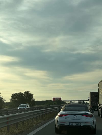 The image captures a view from a car on a highway, likely the A1 highway near Belozem, Bulgaria, given the presence of a KFC billboard. The sky is overcast with scattered clouds. In the foreground, a white Mercedes-Benz convertible is in the right lane, its brake lights illuminated. The license plate is visible: CB 4700 OM. To the left, a silver SUV is in the left lane. A metal guardrail runs along the side of the highway. In the distance, a large KFC billboard is visible on the left, and overhead structures, possibly a bridge or overpass, span the highway. On the right side of the highway, a large truck is partially visible. The environment appears to be a rural or semi-rural area with trees lining the side of the road.