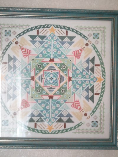 The image shows a framed cross-stitch artwork. The artwork features a circular mandala-like design with a Christmas theme. The design incorporates stylized Christmas trees, angels, and gift boxes in various shades of green, blue, white, and red. The outer border of the artwork has a decorative pattern. The artwork is enclosed in a teal-colored frame with a slightly distressed finish and a gold inner trim. The background of the image appears to be a textured wallpaper. The location of this artwork within the city of Первомайская, Russia cannot be determined from the image alone.