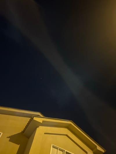A low-angle, nighttime shot captures the exterior of a tan-colored building juxtaposed against a dark sky. A thin band of clouds stretches diagonally across the frame, punctuated by faint stars. The building's architecture features distinct geometric shapes and window details. No people are visible. The scene lacks any specific information to identify the location as La Aurora, Ecuador.