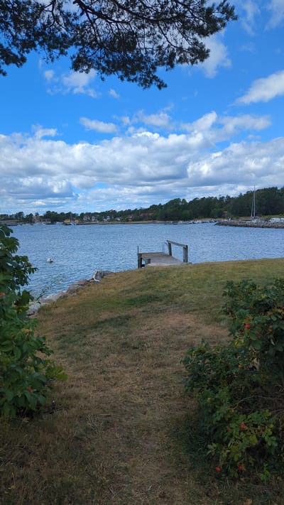 The image depicts a scenic view of the waterfront in Larkollen, Norway. A wooden jetty extends into the calm blue water, with several boats docked along the distant shoreline. The coast is lined with trees and houses, under a sky filled with fluffy white clouds against a bright blue backdrop. In the foreground, a grassy lawn slopes down towards the water, bordered by lush green bushes. The overall scene is tranquil and picturesque, capturing the essence of a serene coastal environment.