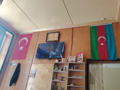 The image depicts an indoor scene, likely in a residential or small business setting, in the city of Khudat, Azerbaijan. Dominating the wall are two national flags: the Turkish flag on the left and the Azerbaijani flag on the right. A flat-screen television is mounted on the wall, displaying a program. Below the television, a white shelf unit is filled with various snack packages and other items. A black jacket hangs from a coat rack on the wall. A framed picture, possibly a religious or historical depiction, hangs above the television. The ceiling is constructed of white panels, and a small security camera is visible near the ceiling. The overall impression is one of a modestly decorated and functional space.