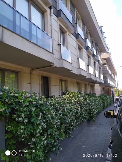 An outdoor, eye-level, slightly wide shot captures the facade of a modern, multi-story apartment building with a row of flowering green bushes in the foreground. The building, painted a light beige, features multiple balconies with glass railings, set against a bright, overcast sky. The bushes are dense and lush, with small white flowers scattered throughout. A paved walkway runs alongside the bushes, and a dark-colored car is partially visible on the right side of the frame. The timestamp "2026/6/18 17:52" is overlaid on the lower right of the image, and "REDMI NOTE 7 ТУРЦИЯ" is in the lower left. The scene appears to be in the Avcılar district of Istanbul, Turkey, given the "ТУРЦИЯ" text. There are no people or animals visible in the frame. The overall impression is one of a well-maintained urban residential setting.