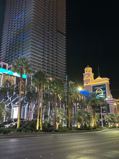 A nighttime view of the Las Vegas strip featuring the Aria hotel on the left, recognizable by its unique geometric design with blue accent lights. The Bellagio hotel and its iconic sign, promoting "O" by Cirque du Soleil, is visible further along the strip. Palm trees line the street, illuminated with string lights, creating a warm glow. The street is visible on the right side of the image.