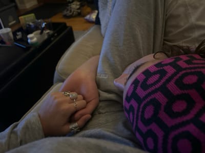 The image captures an intimate indoor scene featuring two individuals. One individual, partially visible in the foreground, is resting or asleep, positioned on their side with closed eyes. They wear a magenta and black headband with a repeating geometric pattern and a grey top. Dark hair is visible near the headband. Adjacent, the hand of a second individual holds the resting person's hand. This hand, also wearing grey fabric on the arm, features three rings: one on the ring finger with a light, rounded cabochon stone, a wavy silver ring on the middle finger, and a textured silver ring on the pinky finger. Both individuals appear to be wearing casual, grey clothing. In the blurred background, a dark, flat surface or table holds several indistinct objects, including a white disposable cup, a white plastic bottle, and other small items. A light-colored upholstered surface, likely a couch, supports the individuals. Further back, a wooden floor and additional blurred household objects are visible. The lighting is subdued. Based on the visual evidence, the specific location within Kingston, Canada, cannot be determined.