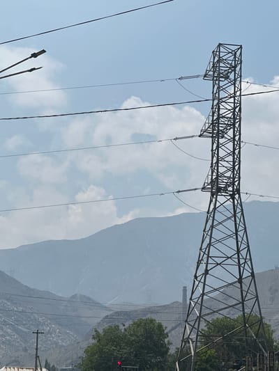 A tall, lattice-style electricity pylon dominates the right side of the frame, its metallic structure silhouetted against a hazy blue sky dotted with soft white clouds. Power lines stretch horizontally across the sky, intersecting with the pylon. In the distance, hazy mountain ranges recede into the background. Below the mountains, a more distant, slender industrial chimney stands out against the muted landscape. Closer to the foreground, lush green trees partially obscure the lower section of the pylon. A traffic light, displaying a red signal, is visible near the bottom center of the image. Electrical poles and more power lines are scattered across the scene. The overall impression is a juxtaposition of industrial infrastructure and natural landscape, set under a partly cloudy sky.